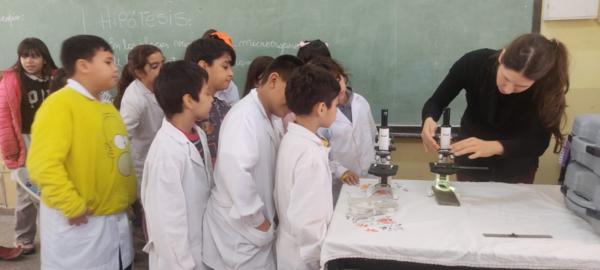 Schoolchildren in white lab coats observe Silvina Alessio from ReAct Latin America as she demonstrates a microscope and teaching about the importance of microorganisms in soil health and food production.