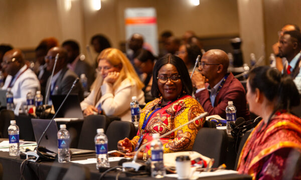 A medium shot of conference attendees seated at tables, focusing on a smiling woman wearing glasses and a colorful patterned top conversing with another participant during a session.