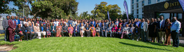 A large group of nearly 200 conference participants and delegates standing and seated outside on a green lawn in front of trees and a building at the ReAct Africa Annual AMR Conference 2026 in Lusaka, Zambia.