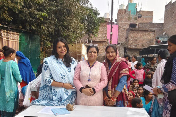Three organizers standing outdoors in a residential neighborhood behind a registration table, displaying the logos of ReAct Asia Pacific and Team of Ek Jeevan Hamari Prerna. Community members, including women and children, gather around the event.