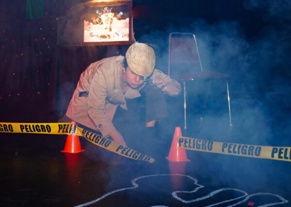 A woman dressed as a detective crouches beside yellow danger tape on a dark stage, examining a marked floor near orange traffic cones.