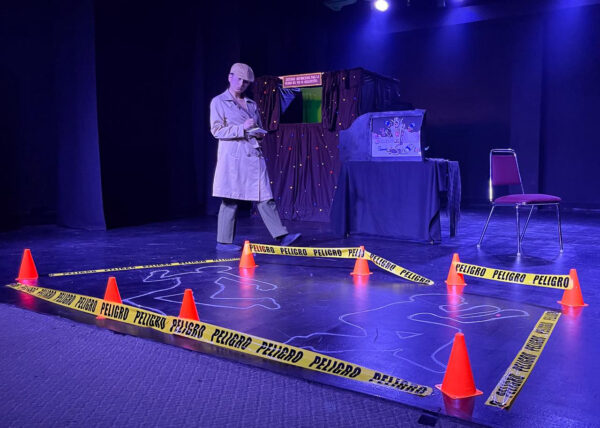 A performer dressed as a detective stands on a dimly lit stage beside a screen, with yellow danger tape and orange cones marking a simulated crime scene.