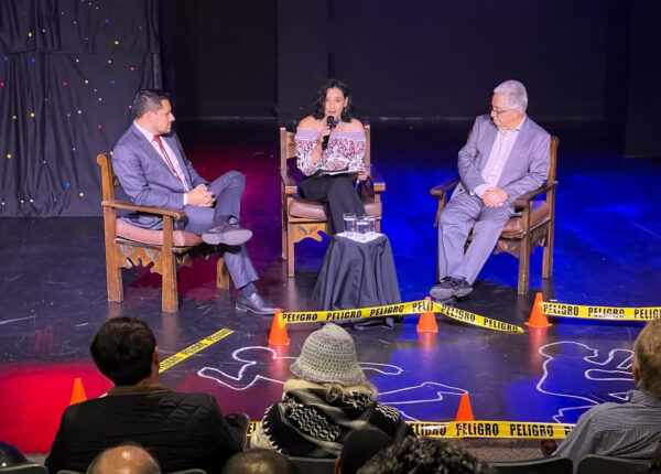 Three people seated on stage during a discussion, with a woman holding a microphone between two male speakers and audience members visible in the foreground.