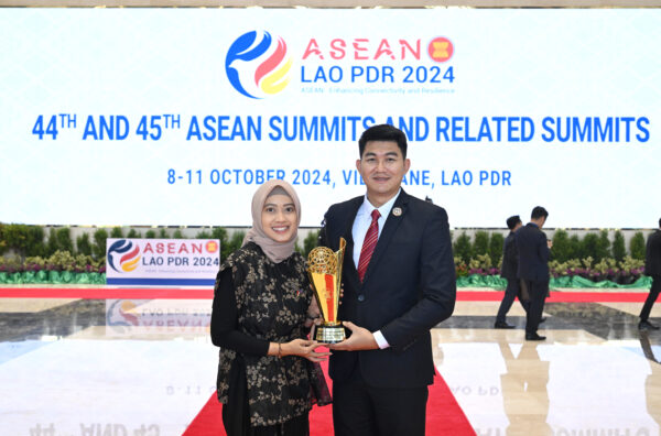 A young woman wearing a headscarf and a young man in a suit stand together holding a trophy at an ASEAN event in Vientiane, Lao PDR. The backdrop identifies the 44th and 45th ASEAN Summits and related summits, held in October 2024.