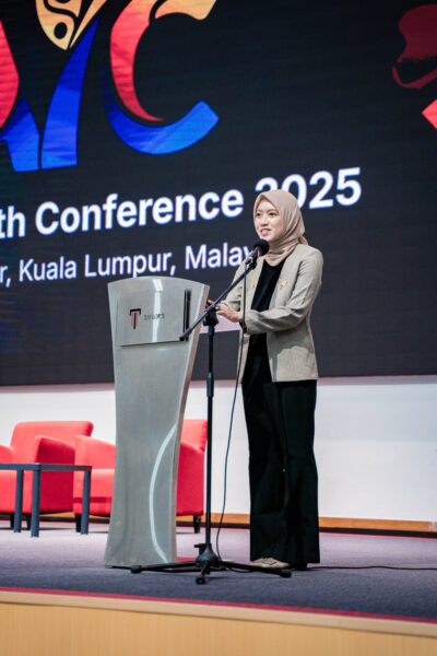 A young woman wearing a headscarf and grey blazer speaks at a podium on a conference stage. A large screen behind her displays text referring to a 2025 conference in Kuala Lumpur.