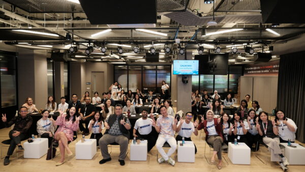 A large group of young people pose together in a conference room, smiling and making peace signs and other gestures. A screen behind them displays “Learn and Play Day.”