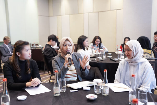 A group of young people, including Sarah Rauzana in a beige headscarf and grey blazer, gather closely for a group selfie at a conference. A participant in the foreground holds up a smartphone to take the photograph.